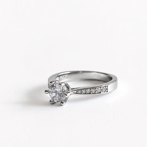 A silver-toned solitaire engagement ring with a round-cut center stone and a pavé-set band, shown on a plain white background.