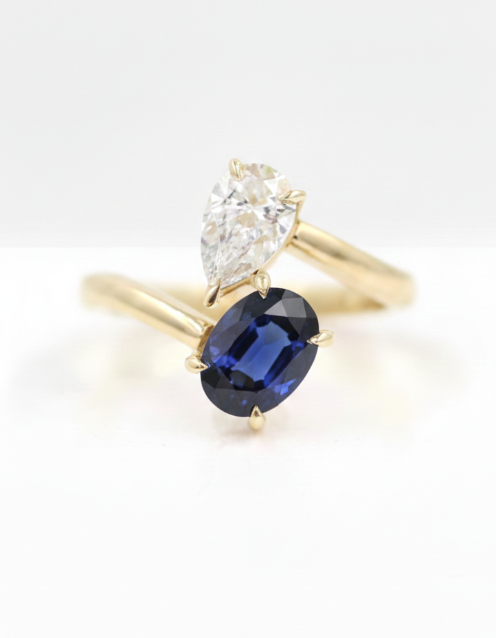 High-resolution macro jewelry photography of a "Toi et Moi" bypass ring. Featuring a deep royal blue oval-cut lab sapphire paired with a brilliant pear-cut moissanite.