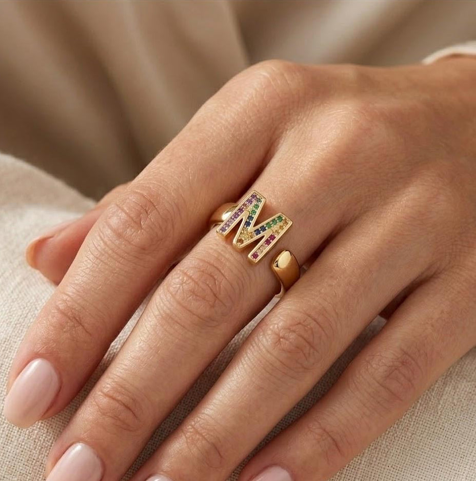 A close-up of a hand wearing a gold ring with colorful gemstone accents and a small heart-shaped detail on the band, resting on a soft beige fabric surface.