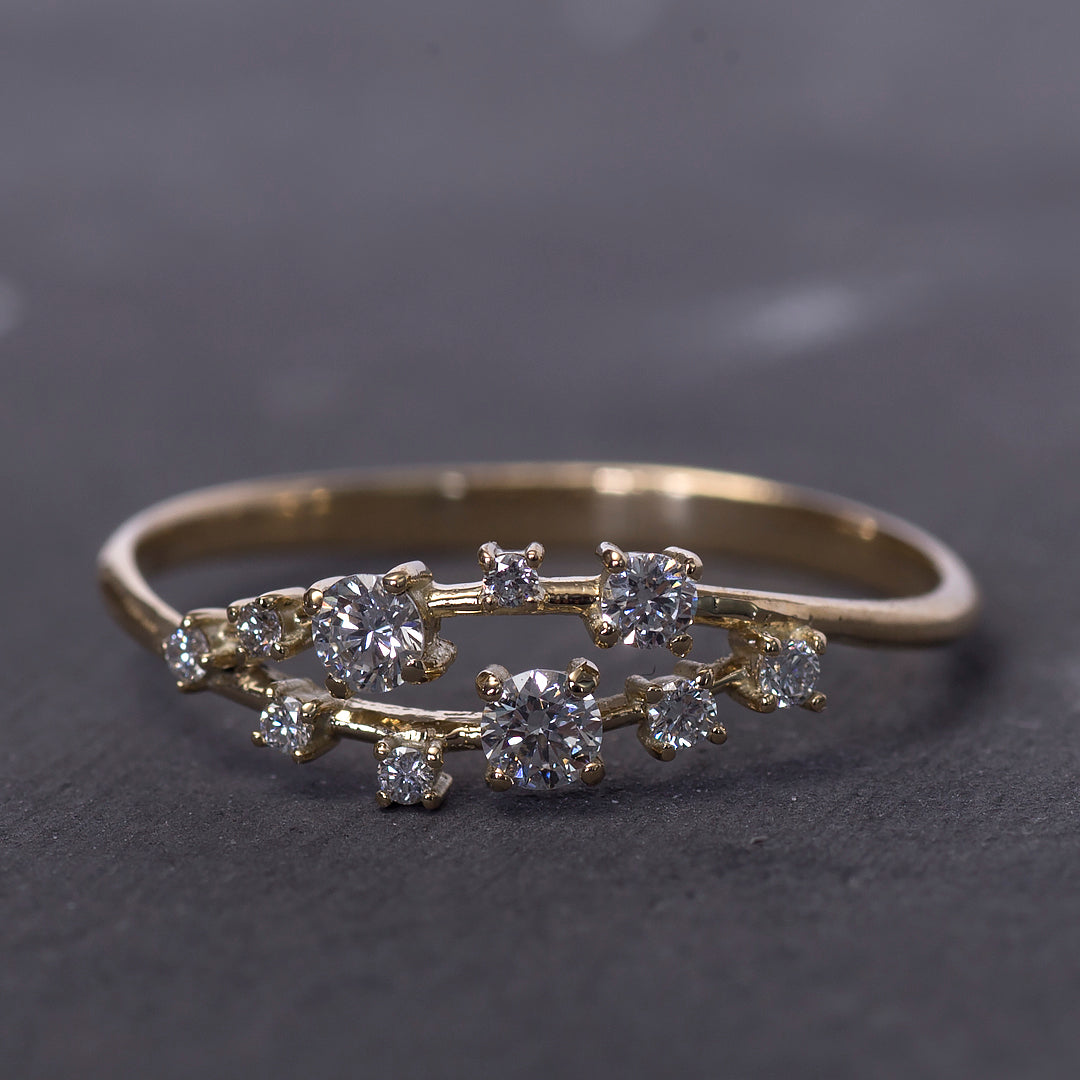 The Diamond Cluster Ring features a delicate 14K gold double band, accented with scattered small round diamonds, and is displayed on a dark textured surface.