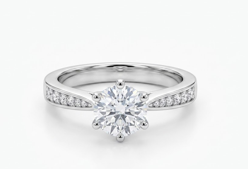 A silver-toned diamond engagement ring with a round-cut center stone and small pavé diamonds along the shoulders, shown on a clean white background.