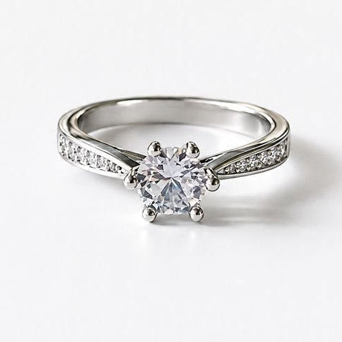 A silver-toned solitaire engagement ring with a round-cut center stone and small pavé accents on the band, shown on a plain white background.