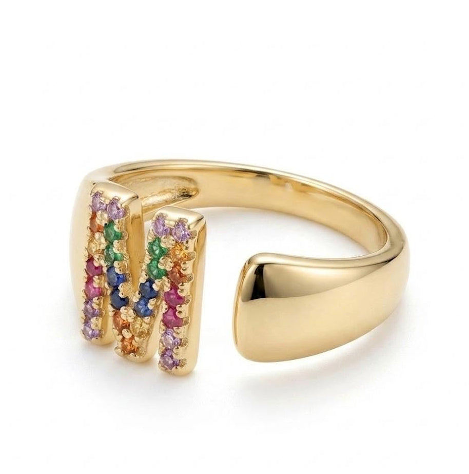 A gold-plated open band ring featuring a large letter 'M' adorned with small multicolored gemstones, shown on a clean white background.